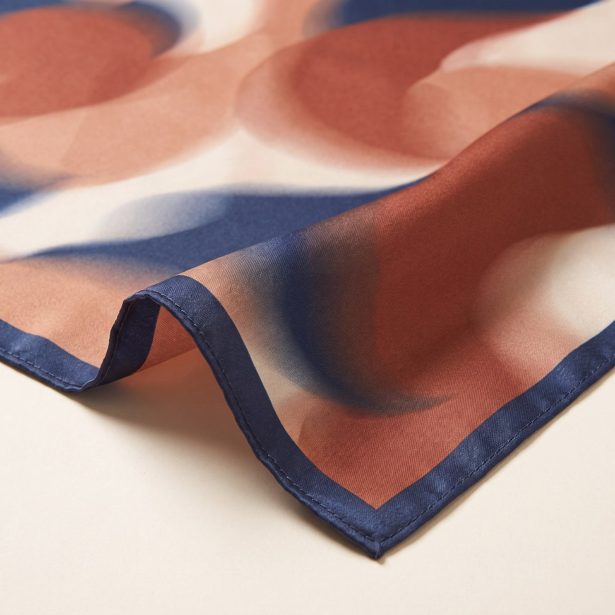 Close-up of the stitched hem, navy border and abstract print of the square satin scarf